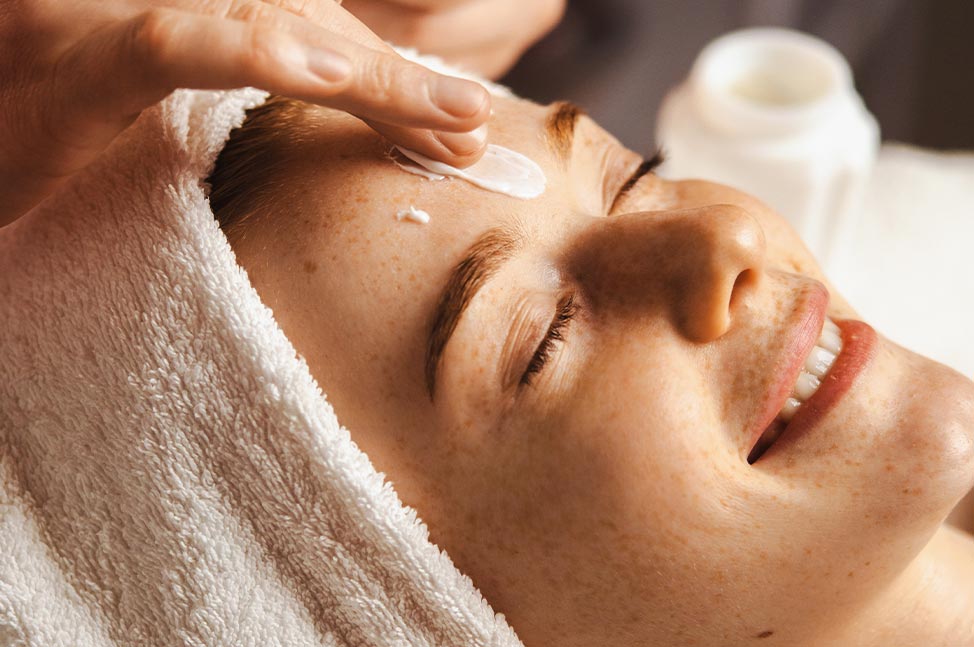 Femme souriante les yeux fermés pendant l’application d’une crème de soin sur le visage.